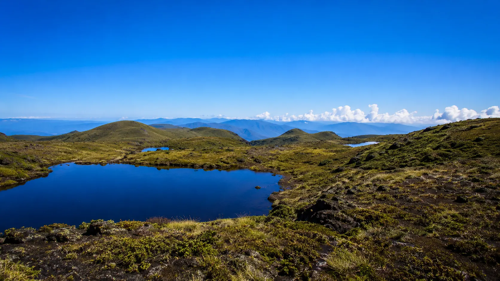 Laguna de agua azul oscura en el páramo del Chirripó, rodeada de cerros redondeados cubiertos de pasto y roca negra, bajo un cielo azul intenso.